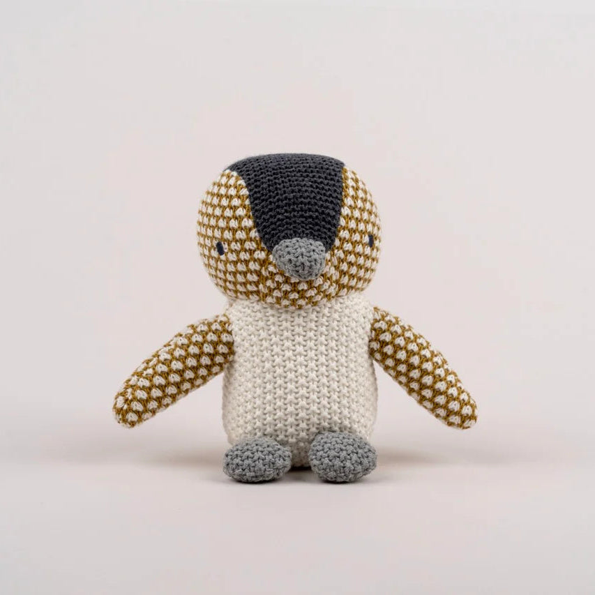 A handmade crocheted penguin toy with a cream-colored body, brown and white patterned arms and head, and gray feet and beak, set against a plain light gray background.