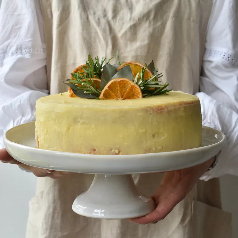 Person holding a lemon cake with orange slices and rosemary on a white plate.