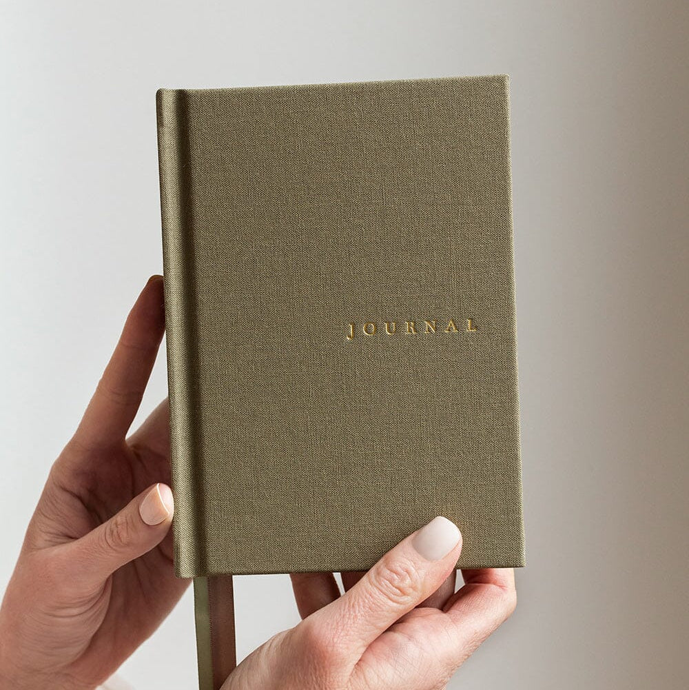 Hand holding a green journal with 'JOURNAL' embossed on it against a light background
