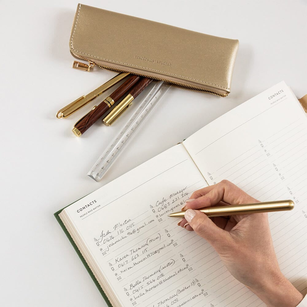 Person writing in a notebook with a gold pen, surrounded by stationery items on a light surface.