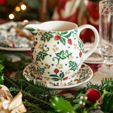 A white ceramic Christmas creamer pitcher with a green holly and red berry pattern, resting on a matching saucer on a decorated holiday table with pine branches and warm blurred lights in the background.