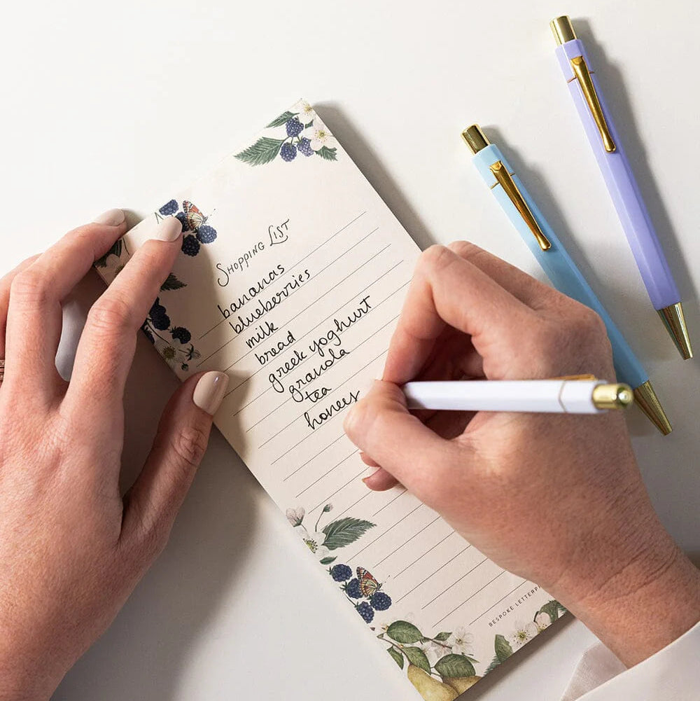 Person writing in a floral notebook with three pens on a white surface