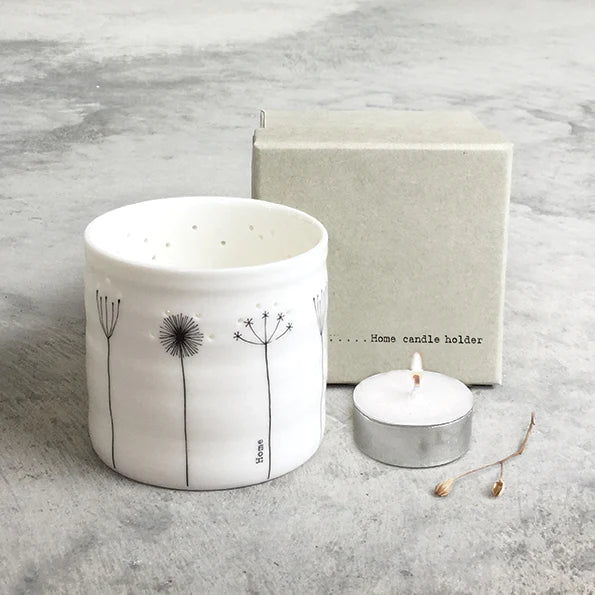 A white porcelain tea light holder with a clear glaze, featuring black line illustrations of plants and the word 'Home'. It is displayed next to a lit tea light and a box, on a grey background.