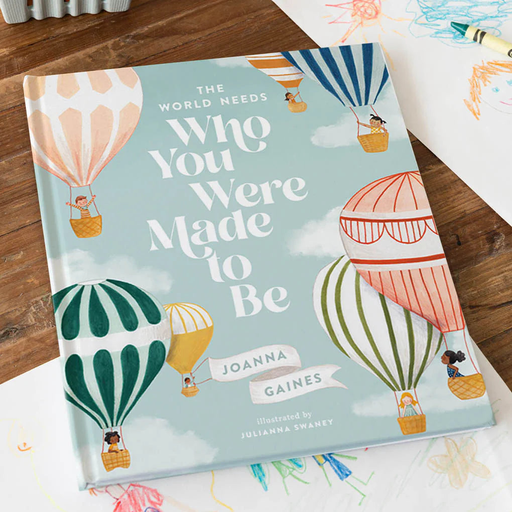 Book titled 'The World Needs Who You Were Made to Be' by Joanna Gaines on a wooden surface with colorful illustrations of hot air balloons.