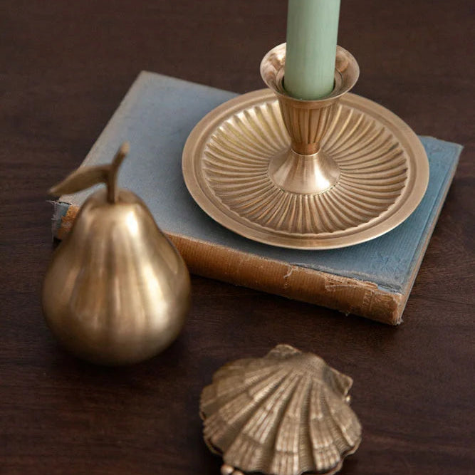 A brass candlestick holder with a light green candle, styled on top of a vintage blue book alongside a brass pear and a brass shell on a dark wooden table.