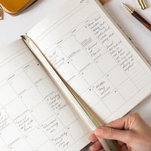 Open planner with handwritten notes on a white surface, held by a hand with a pen.
