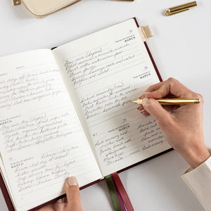 Person writing in planner with gold pen on a white background.