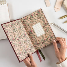 Floral-patterned notebook held by hands on a white surface with stationery items.