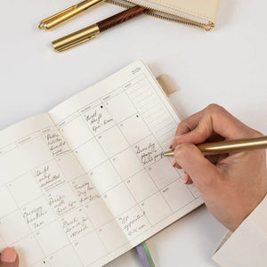 Person writing in a planner with a gold pen on a white surface