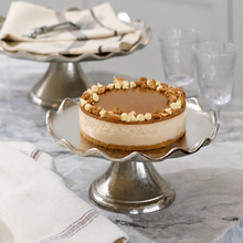 A round cheesecake with a glossy caramel-like topping and white chocolate chips sits on a silver pedestal cake stand on a marble countertop. A folded cream cloth with a fork is behind it, and two blurred drinking glasses are in the background.