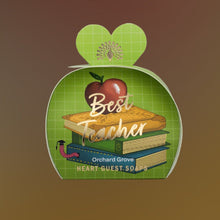 A decorative gift box or packaging insert shaped like a green apple with a heart-shaped top, featuring an apple, books, and the text 'Best Teacher' and 'Orchard Grove HEART GUEST SOAPS' on a brown gradient background.