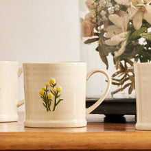 White mug with floral design on a wooden surface, with a vase of flowers in the background.