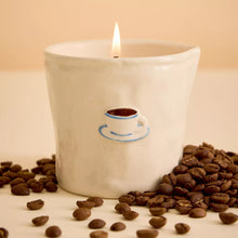 A white ceramic candle with a small coffee cup and blue saucer detail, lit with a flame and surrounded by scattered coffee beans on a warm beige surface and background.