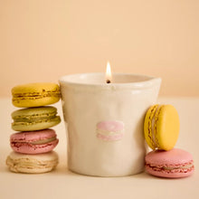 A white ceramic candle holder with a lit flame, decorated with a small pink macaron illustration, styled with colorful macaron stacks on a warm beige background.