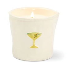 A lit cream-colored ceramic candle in a small cup-shaped vessel with a painted green cocktail glass design on the front, set against a plain white background.