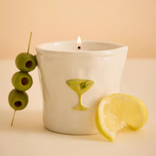 A white ceramic candle holder with a lit candle, decorated with a green martini glass motif and green olives on a skewer, styled with a lemon slice against a warm beige background.