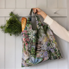 A reusable fabric tote bag with a vintage floral and botanical print is hanging on a white paneled door. A hand wearing a cream-colored long-sleeve blouse holds the bag, which also contains a loaf of bread and a bunch of fresh dill.