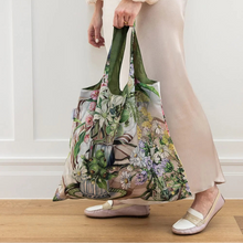 A person wearing a cream floral print blouse and beige satin skirt holds a large, reusable tote bag with a green handle and a multicolor botanical print. The person is standing on a light wood floor against a white paneled wall.