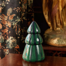 A glossy, tiered Christmas tree-shaped ceramic candle in green and navy stripes, displayed on a dark wooden table with a floral curtain and a gold statue in the background.