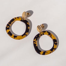A pair of large open circular drop earrings with a yellow and black mottled pattern, attached to gold-tone posts, displayed on a clean white background.