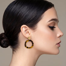 A close-up side profile of a woman with dark hair in a bun, wearing a large circular drop earring with a yellow and black tortoiseshell pattern and a gold stud.