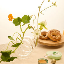 Clear glass vase with green leaves and flowers on a wooden surface with bagels and tea.