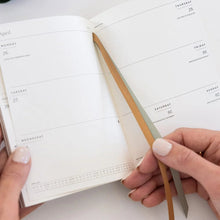 An open weekly planner with white pages and minimalist gray text, held by two hands. A thin dual-tone ribbon bookmark in beige and gray is visible, along with a dark green zip case and gold pen in the background on a white surface.