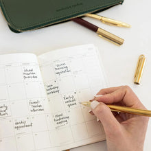 A hand writing in an open monthly planner with a gold pen, with a green leather zip pouch, gold pens, and pen parts arranged on a white surface.