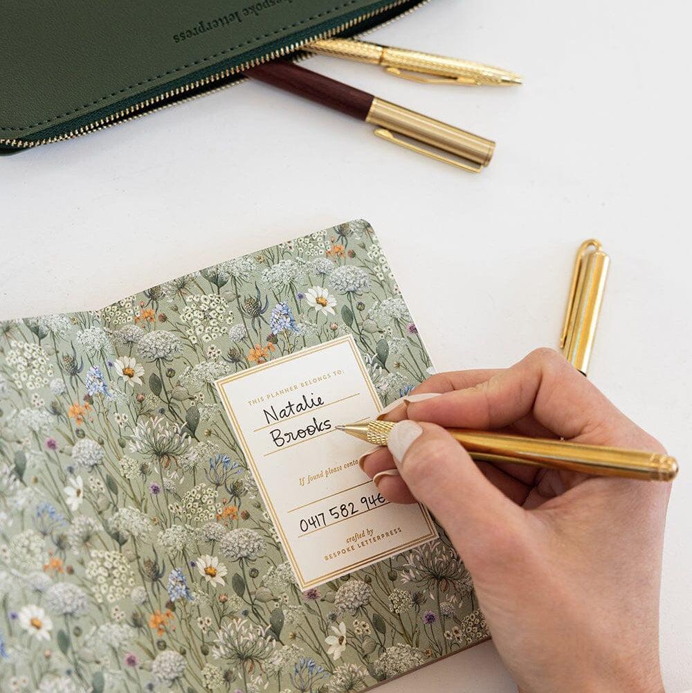 A hand writes in a floral-patterned notebook with a gold pen, with a dark green leather zip pouch and two gold pens on a white surface.