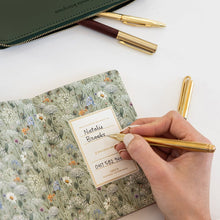 A hand writes in a floral-patterned notebook with a gold pen, with a dark green leather zip pouch and two gold pens on a white surface.
