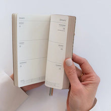 A person's hands holding an open planner showing the interior layout with weekly columns for Monday through Sunday and blank page sections for January 16 and 17, against a clean white background.
