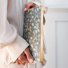 A person in a flowing cream dress holds a floral patterned 2027 journal with gold foil numbers. The background is a clean, bright white interior.
