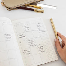 An open planner with a weekly calendar layout on a white surface, alongside a hand holding a gold pen, a gold pencil case, and two gold and brown pens.
