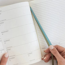 An open weekly planner with lined pages, held by two hands. A slim, dual-colored pen or bookmark with a teal top and olive green bottom rests across the pages.