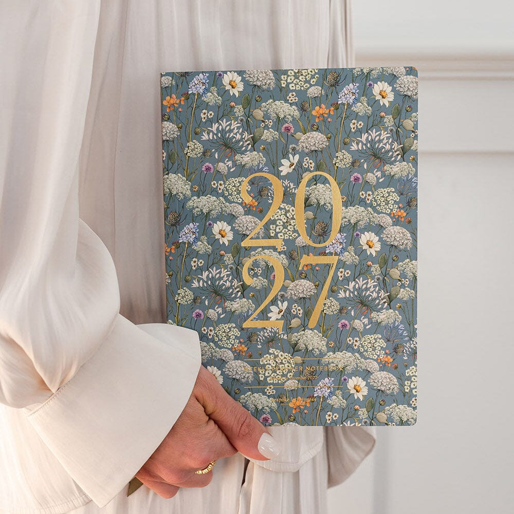 A person in a white dress holds a 2027 floral calendar. The calendar cover features a detailed blue-gray wildflower pattern with large gold '2027' text in the center.