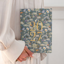 A person in a white dress holds a 2027 floral calendar. The calendar cover features a detailed blue-gray wildflower pattern with large gold '2027' text in the center.