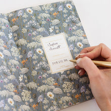 A floral patterned notebook with a blue-gray background and white, yellow, and purple wildflower illustrations, shown open with a hand writing in a gold pen. The cover features a cream label with the text 'Cople Square' and 'Our Size 3.