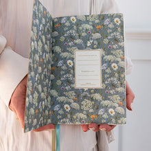 A person in a cream-colored dress holds an open floral journal. The cover and interior are decorated with a detailed pattern of wildflowers in muted blue, white, yellow, and orange tones.
