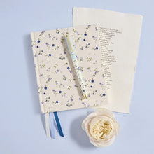 A cream-colored hardcover journal with a delicate blue and green floral pattern, styled with a matching floral pen, an open notebook with handwritten text, and a single cream rose on a light blue background.