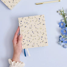 A hand holds a cream-colored notebook with a delicate blue and green floral pattern. The scene is styled on a light blue background with a matching cream pen, a second notebook in the top-left corner, and blue flowers on the right.