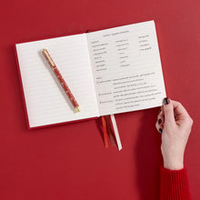 An open lined notebook with handwritten notes on the pages, a red-and-gold pen resting on the left page, and ribbon bookmarks hanging from the bottom. A person's hand with dark nail polish and a red knit sleeve holds the right edge of the notebook against a solid red background.
