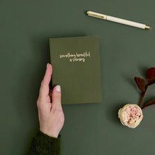 A hand holds an olive green notebook with the handwritten phrase 'something beautiful is coming' on the cover. The scene is styled on a dark green background with a cream pen, a pale pink rose, and a dark green sleeve visible.