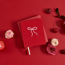 A red hardcover notebook with a white line-art bow design on the cover, styled in a flat lay on a matching red background with decorative pink and red flowers and cherries.