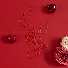 Red metal paperclip desk accessories scattered on a textured red background, styled with two dark red cherries and pink and cream roses.