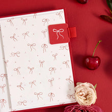 An open notebook or journal with white pages decorated with a repeating red bow pattern, a red cover, and a red bookmark tab with a white bow illustration. The notebook is styled on a textured red background with decorative cherries and flowers.