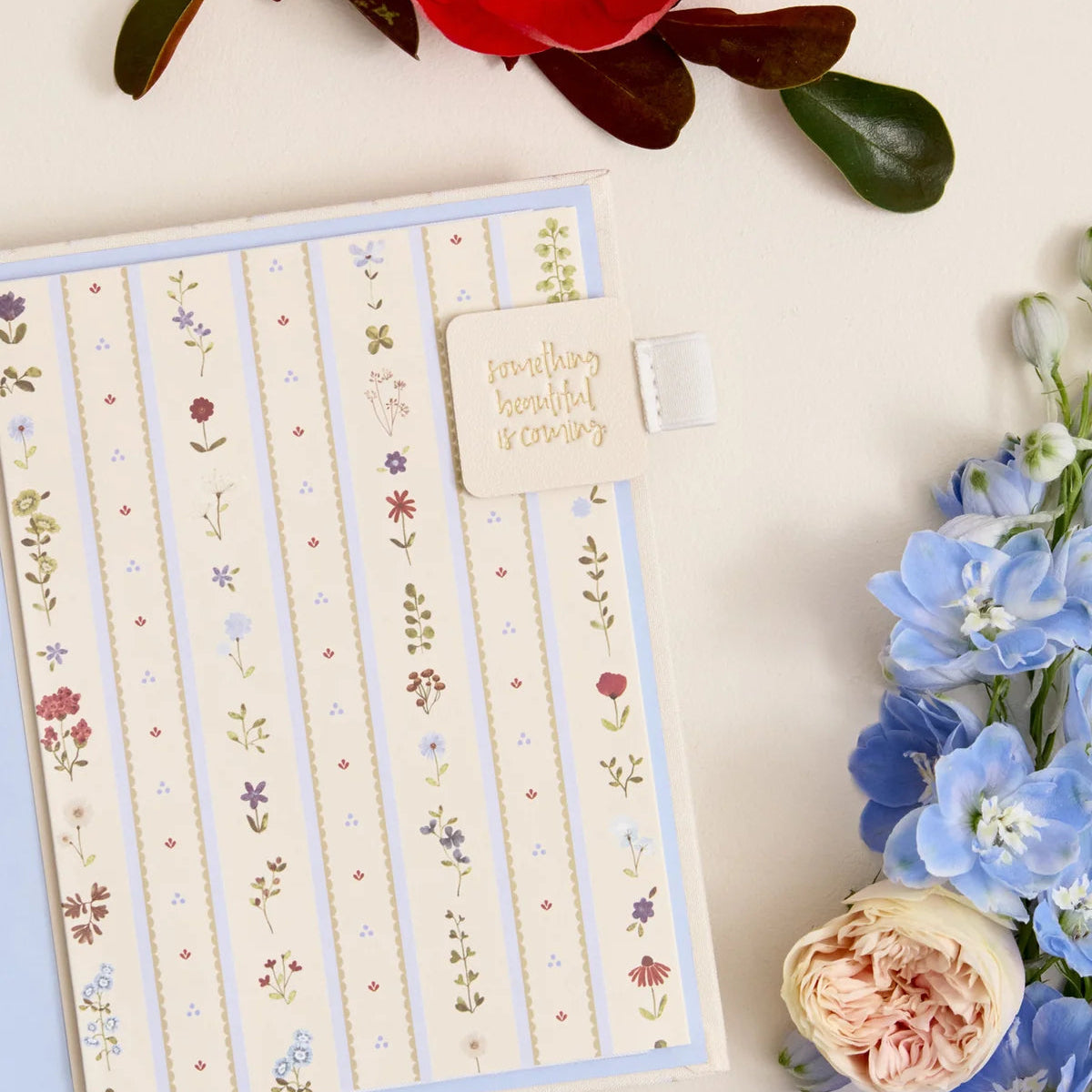 A floral-patterned planner or notebook cover with vertical stripes and small flower illustrations, shown on a cream background with decorative flowers around it. A small tab on the cover reads 'something beautiful is coming.