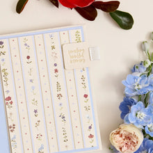 A floral-patterned planner or notebook cover with vertical stripes and small flower illustrations, shown on a cream background with decorative flowers around it. A small tab on the cover reads 'something beautiful is coming.