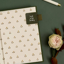 A flat-lay view of a cream-colored notebook with a repeating brown leaf pattern, a dark olive green cover, and a small dark olive green tab on the side with the white script text 'trust in the timing'. A pale pink rose and a white pen with a gold tip are styled around it on a textured dark green background.