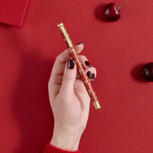 A hand with dark nail polish holds a red and gold decorative pen against a textured red background with cherry props.