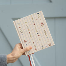 A hand holding a hardcover notebook with a cream-colored cover decorated with vertical stripes and small floral illustrations, set against a light blue-gray paneled background.